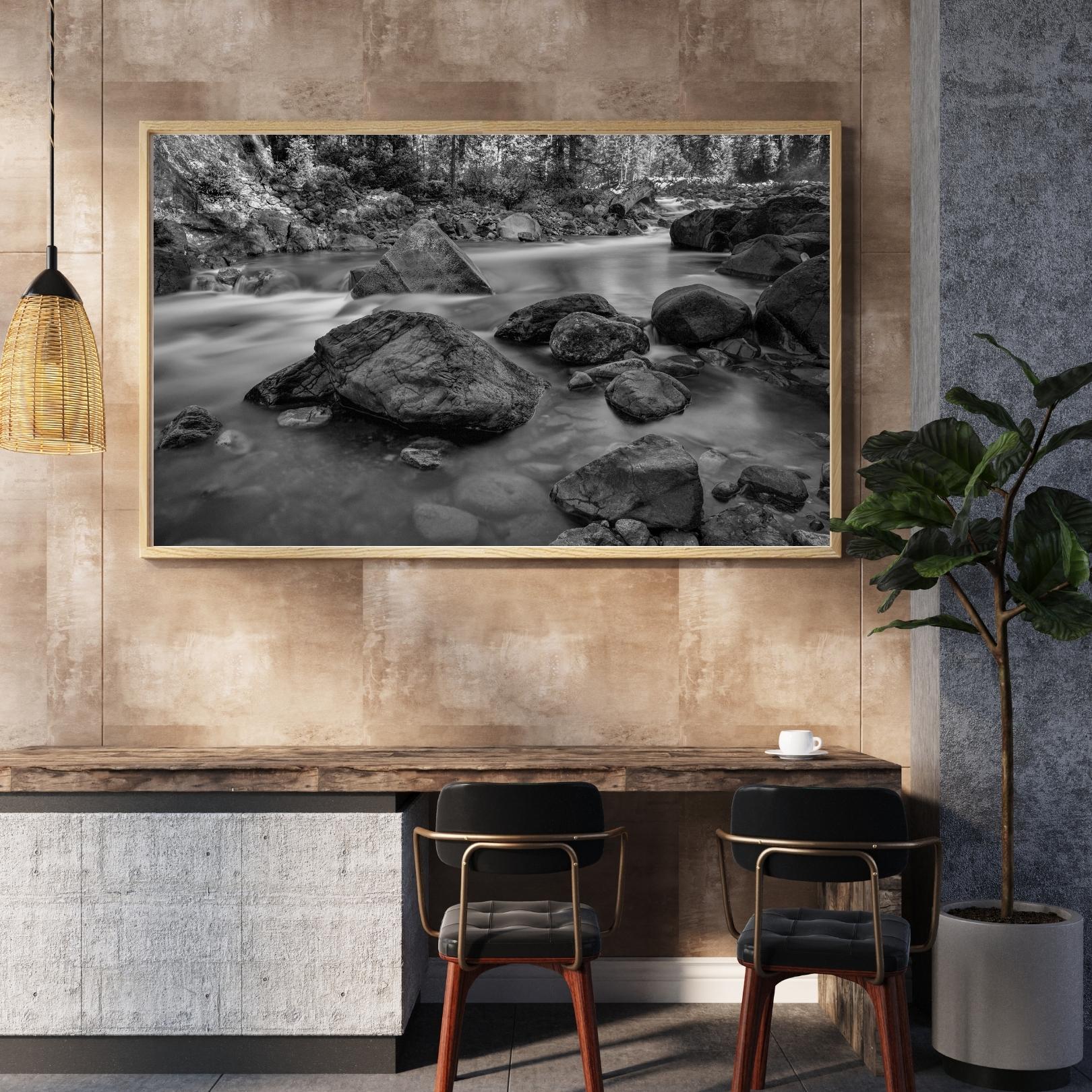 Framed black-and-white photograph of a rocky river scene by Steve Bisig Photography displayed in a modern hospitality workspace with warm textured walls, seating, and contemporary decor.