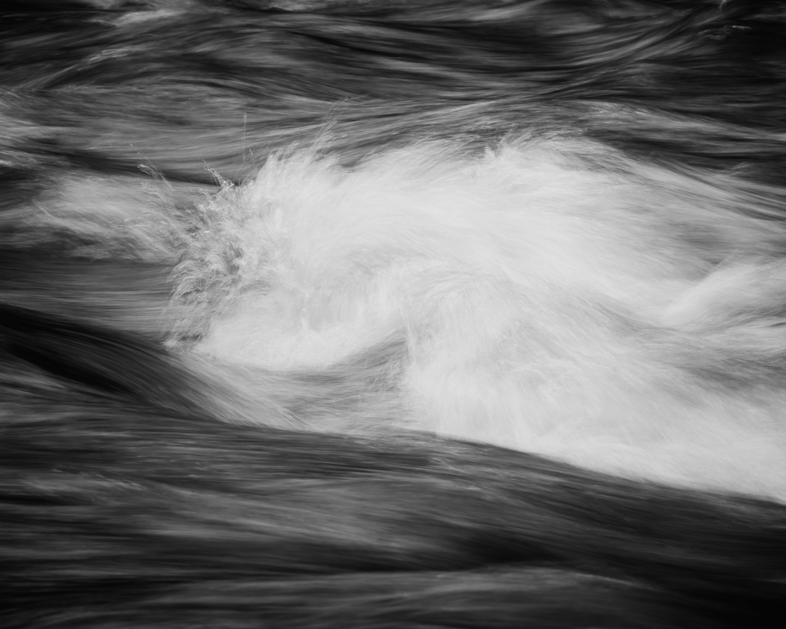 A pale swell of water gathers at the center of the frame, momentarily unified as darker currents stream past and around it.